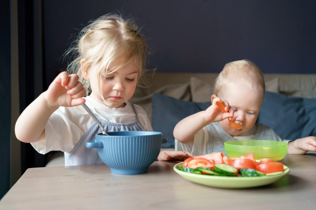Zwei Kinder beim Mittagessen, vor ihnen auf dem Tisch stehen zwei Schüssel und ein Teller. Gesunde Ernährung