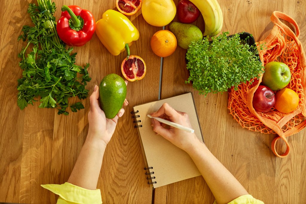 Frau schreibt Speiseplan in ein Notizbuch. Auf dem Tisch liegt frisches Obst und Gemüse für eine gesunde Ernährung