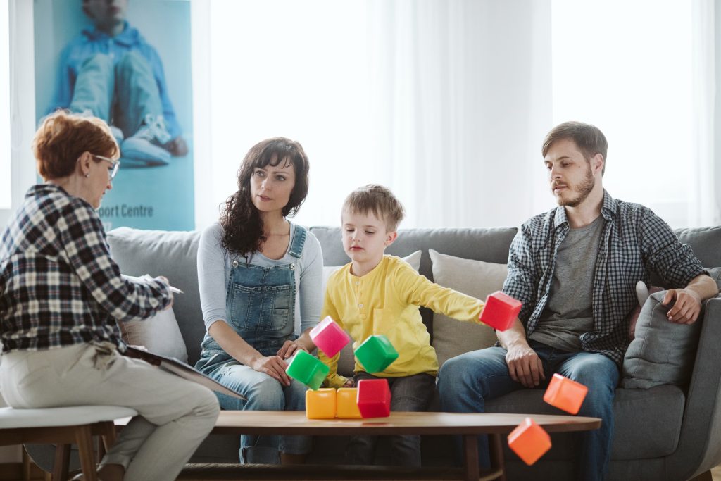 Eine Familie sitzt in einer Beratung, um die Ursachen von Autismus bei ihrem Kind herauszufinden. Der Junge wirft Bausteine vom Tisch. 